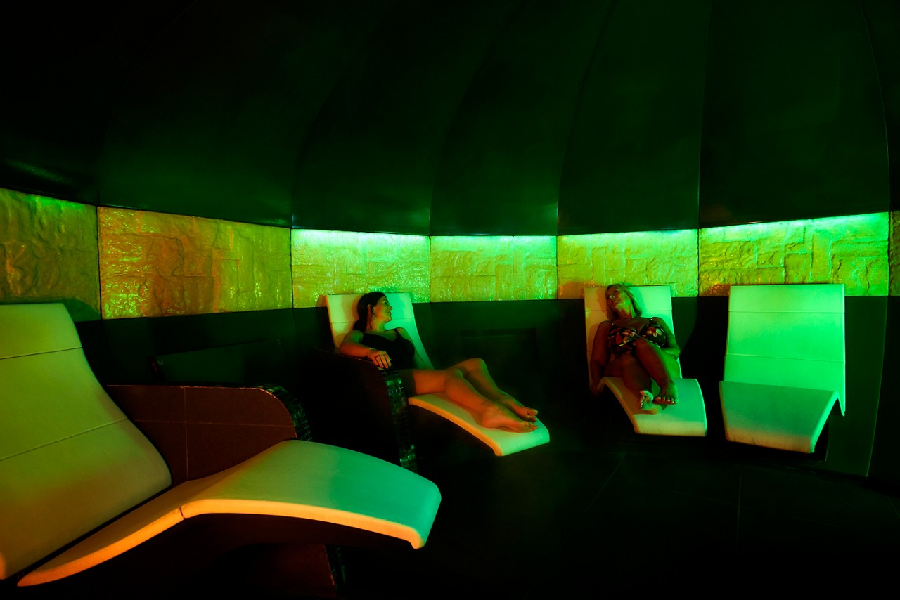 Two people recline on white lounge chairs, relaxing and chatting, surrounded by empty loungers in a dim, green-lit spa room with curved dark walls and a glowing wall band.